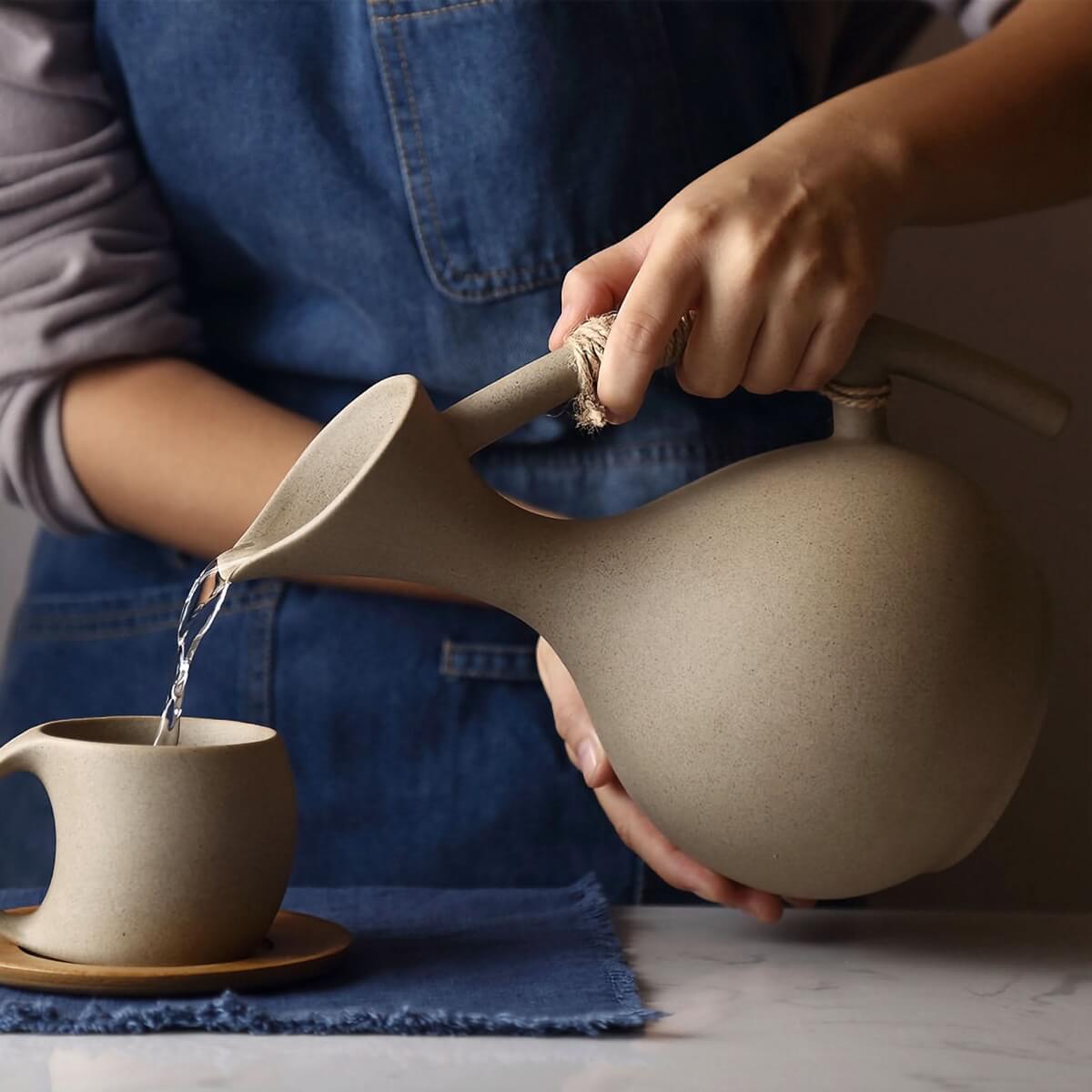 Taupe ceramic pitcher with arched handle pouring water into abstract mug.